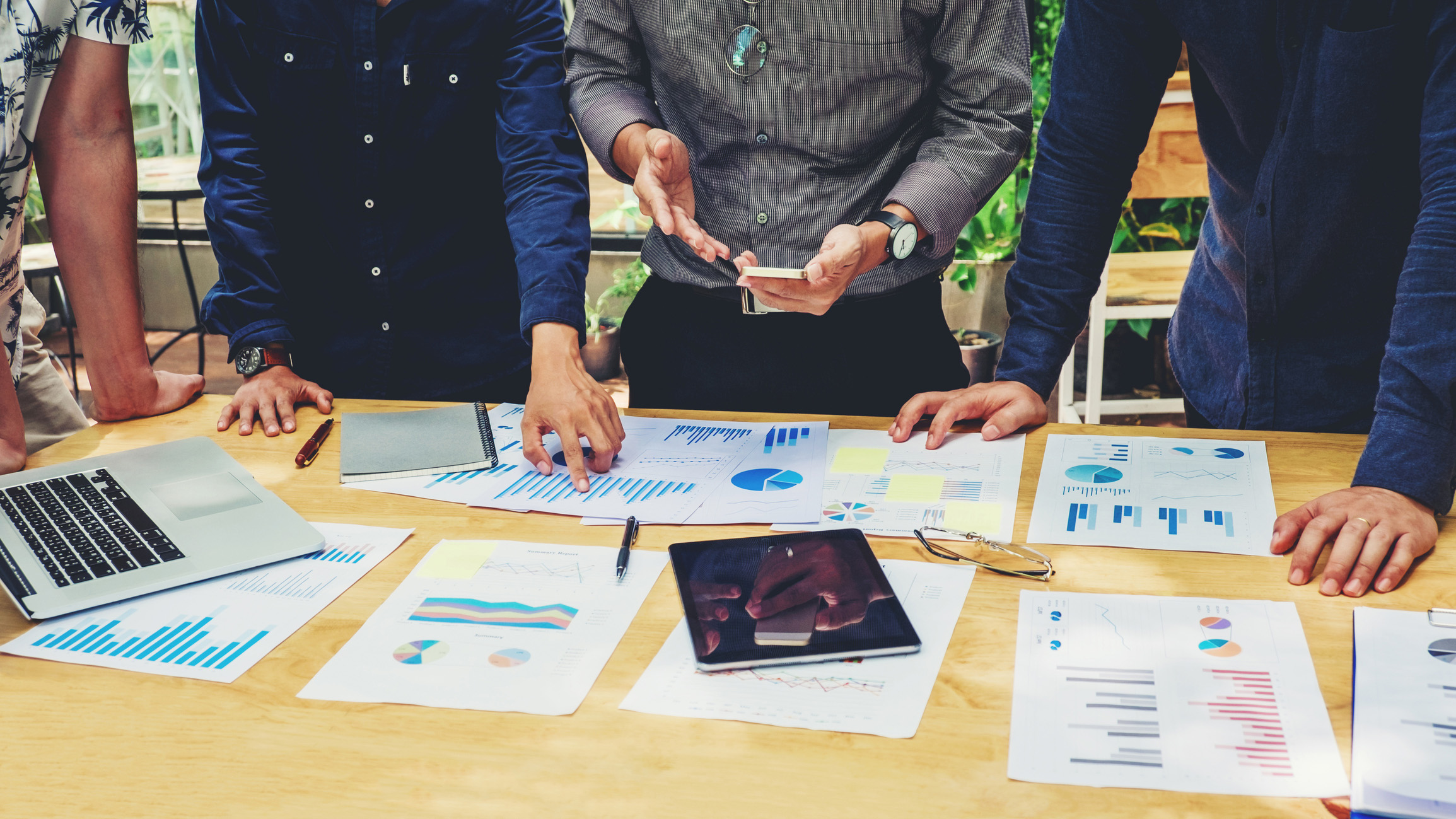 Four people stand around a table covered with charts, graphs, documents, a laptop, and a tablet. One person gestures with their hands while others look at the papers, suggesting a business meeting or discussion.